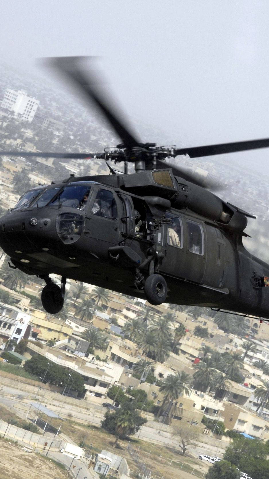 黑色軍用直升機低空飛過有棕櫚樹的城市。