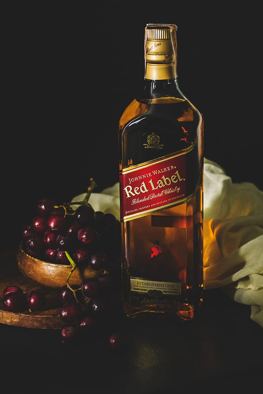 黑麝香葡萄與黑標酒 壁紙 — 一瓶約翰走路紅牌威士忌放在一個木碗旁邊，碗裡裝著深色葡萄，背景為黑色，並有柔軟垂墜的布料。