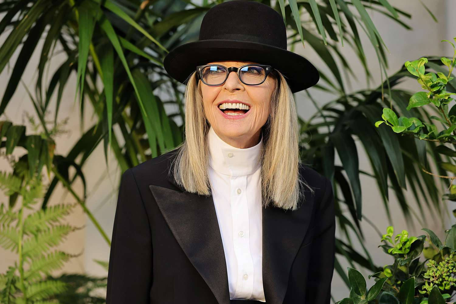 黛安·基頓戴 Ralph Lauren 頭盔帽 壁紙 — 一位金髮戴眼鏡的快樂女性，戴著黑色帽子和西裝外套，在茂密的綠葉前綻放笑容。