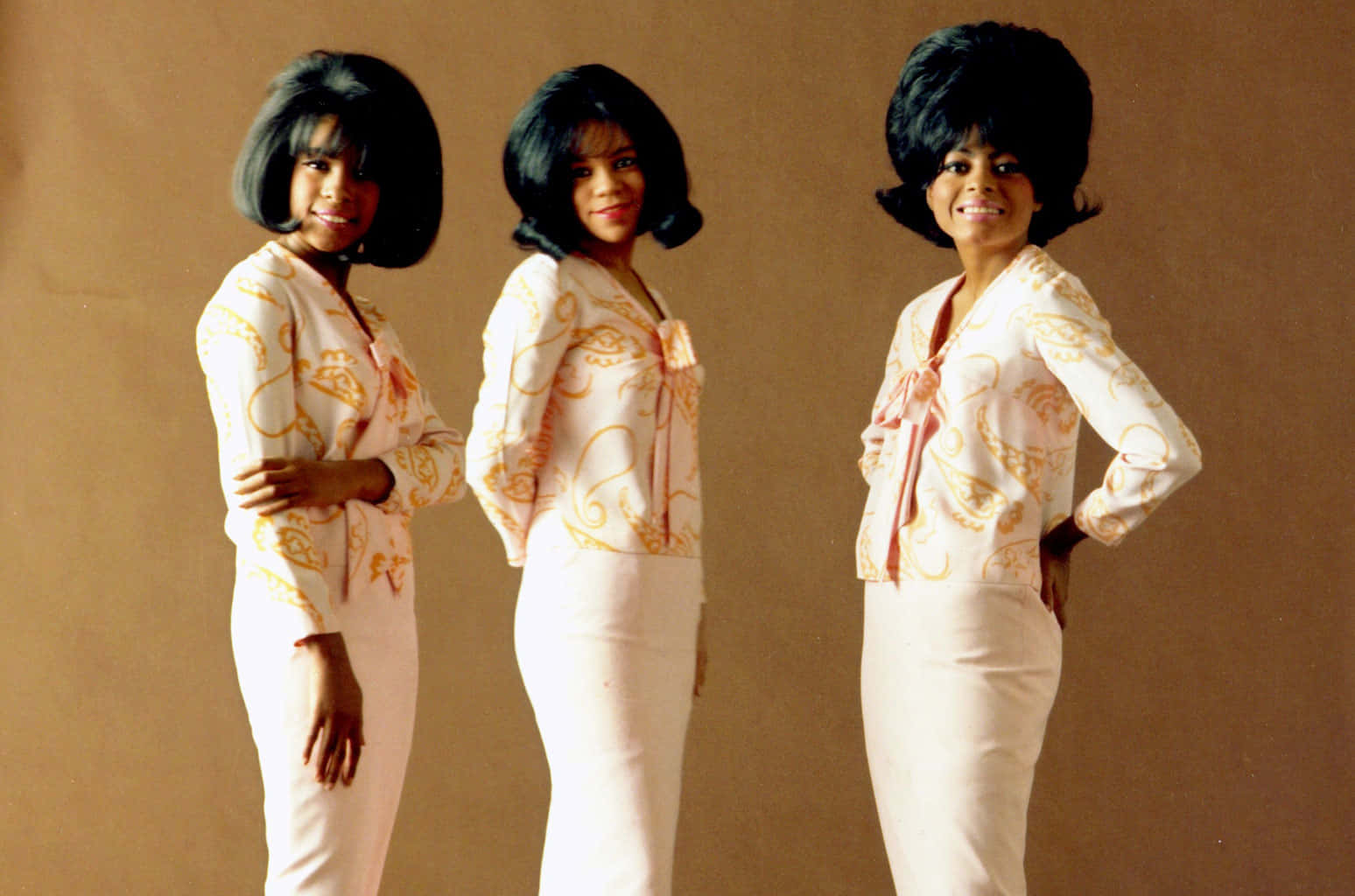 黛安娜·羅斯與 supremes 1964 年彩色肖像 壁紙 — 三位穿著相配白色和金色圖案服裝的女性，在樸素的棕色背景前，以豐盈的黑色髮型自信地擺姿勢。
