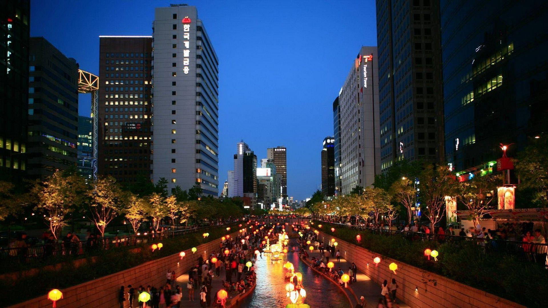 點亮首爾夜空的絢麗燈籠 壁紙 — 黃昏時分繁忙的城市運河，發光的燈籠倒映在水面上，兩旁是高聳的燈火通明的摩天大樓和人群。