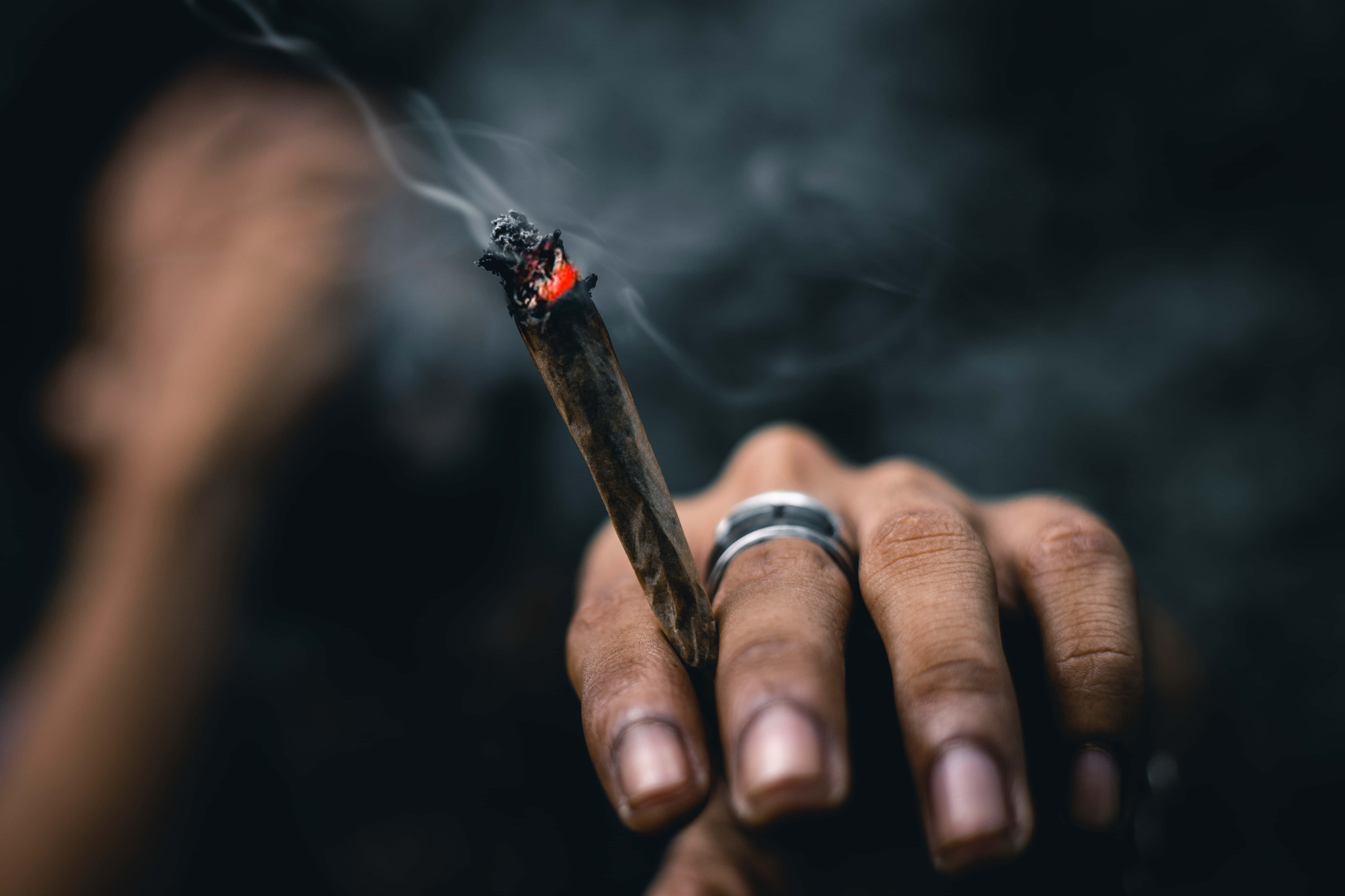 點燃的煙草特寫 壁紙 — 一隻手拿著一支點燃的大麻煙，帶有發光的炭火和繚繞的煙霧，背景是黑暗模糊的。
