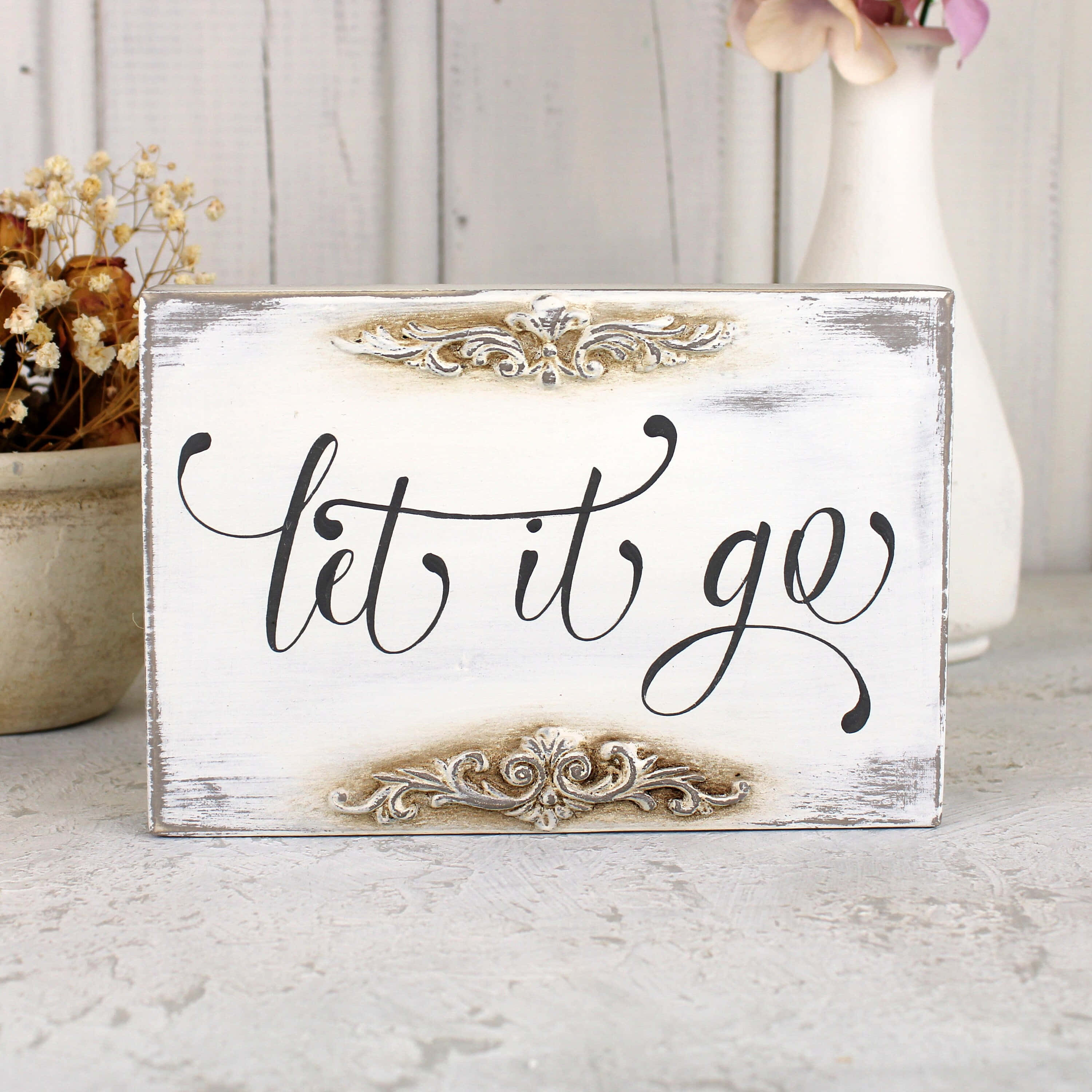 鼓舞人心的「Let It Go」壁畫 壁紙 — 一個破舊的白色木牌，上面有黑色草書文字寫著 let it go，飾有華麗的銀色裝飾，放置在帶紋理的表面上，旁邊有乾花和一個花瓶。