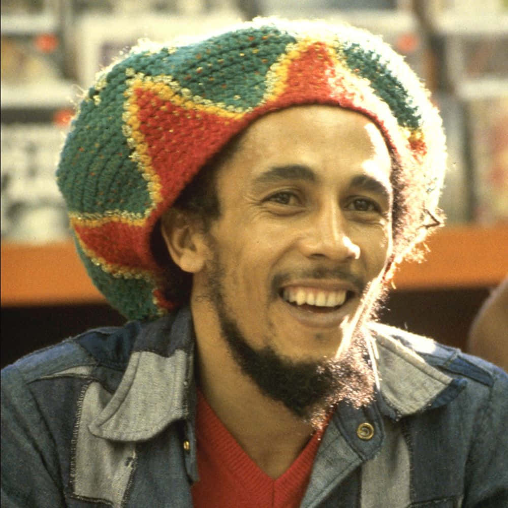 鼓舞人心的雷鬼傳奇人物鮑勃·馬利 壁紙 — Bob Marley 滿懷喜悅，戴著彩色針織帽和牛仔外套，沐浴在溫暖的陽光下。