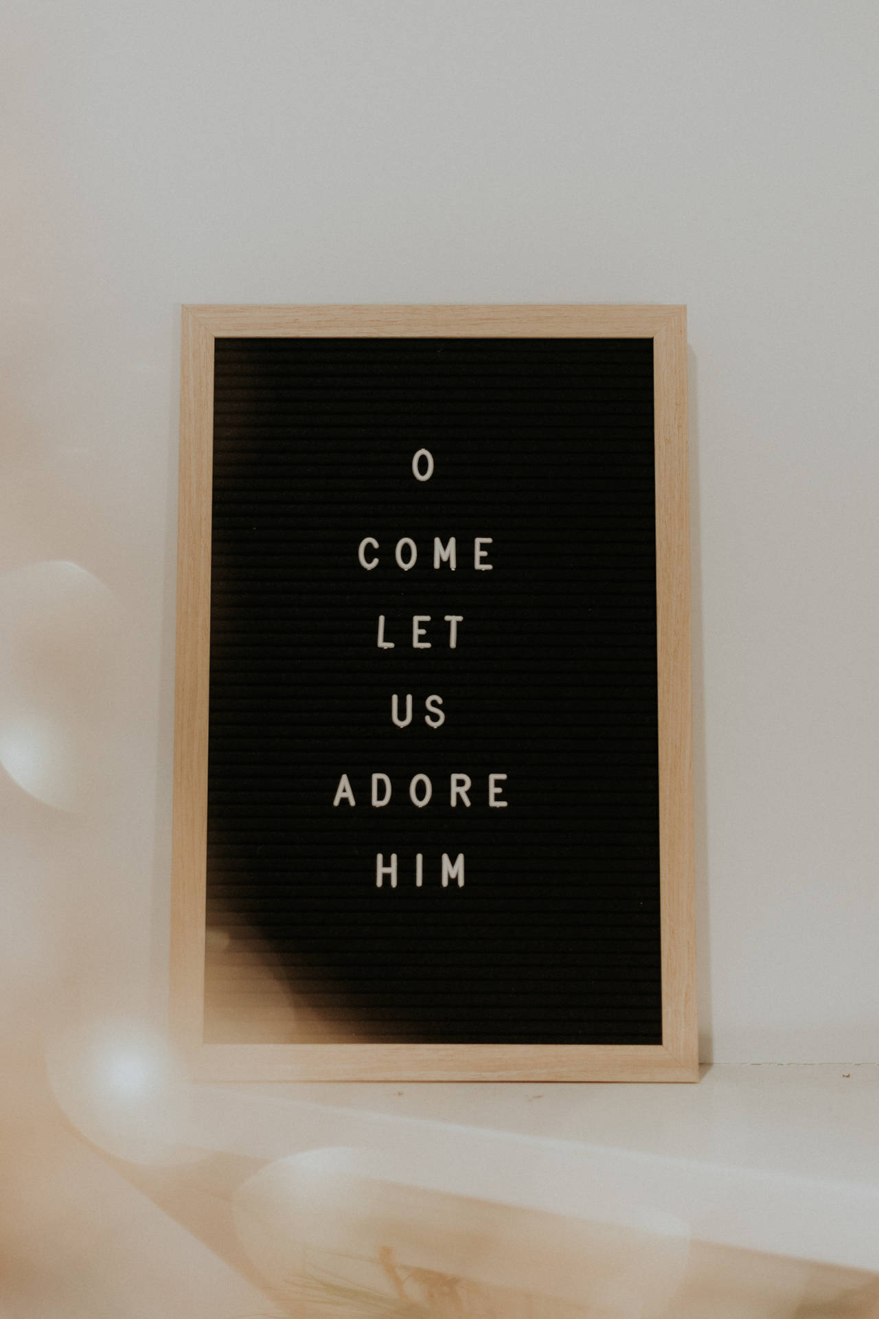 一個木框字母板，白色文字寫著「O COME LET US ADORE HIM」，放在淺色牆上。