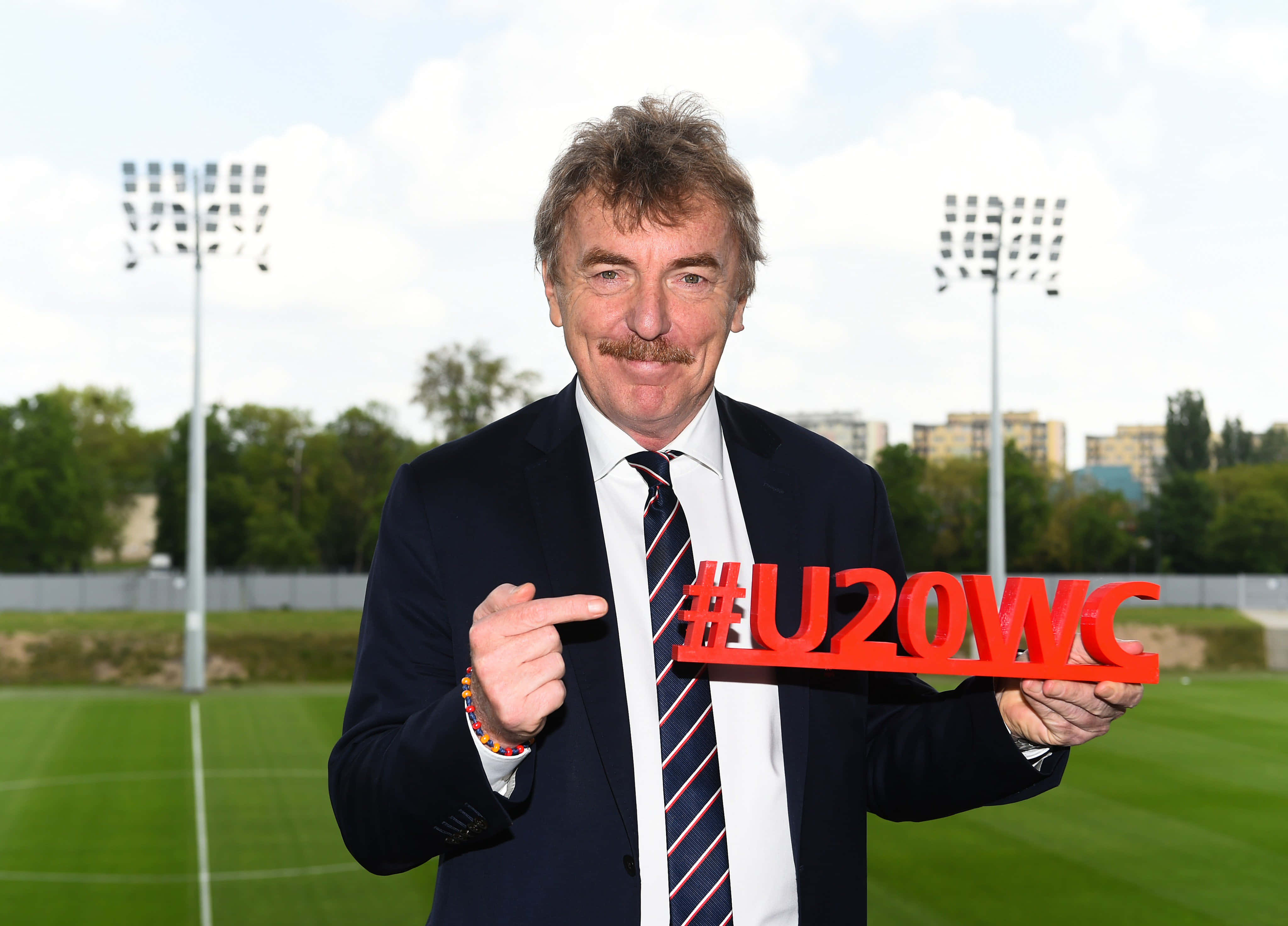 齊格尼·博尼克支持FIFA U20世界盃 壁紙 — 一名微笑的西裝男子在陰天下，於綠色運動場上舉著一個紅色標籤牌。