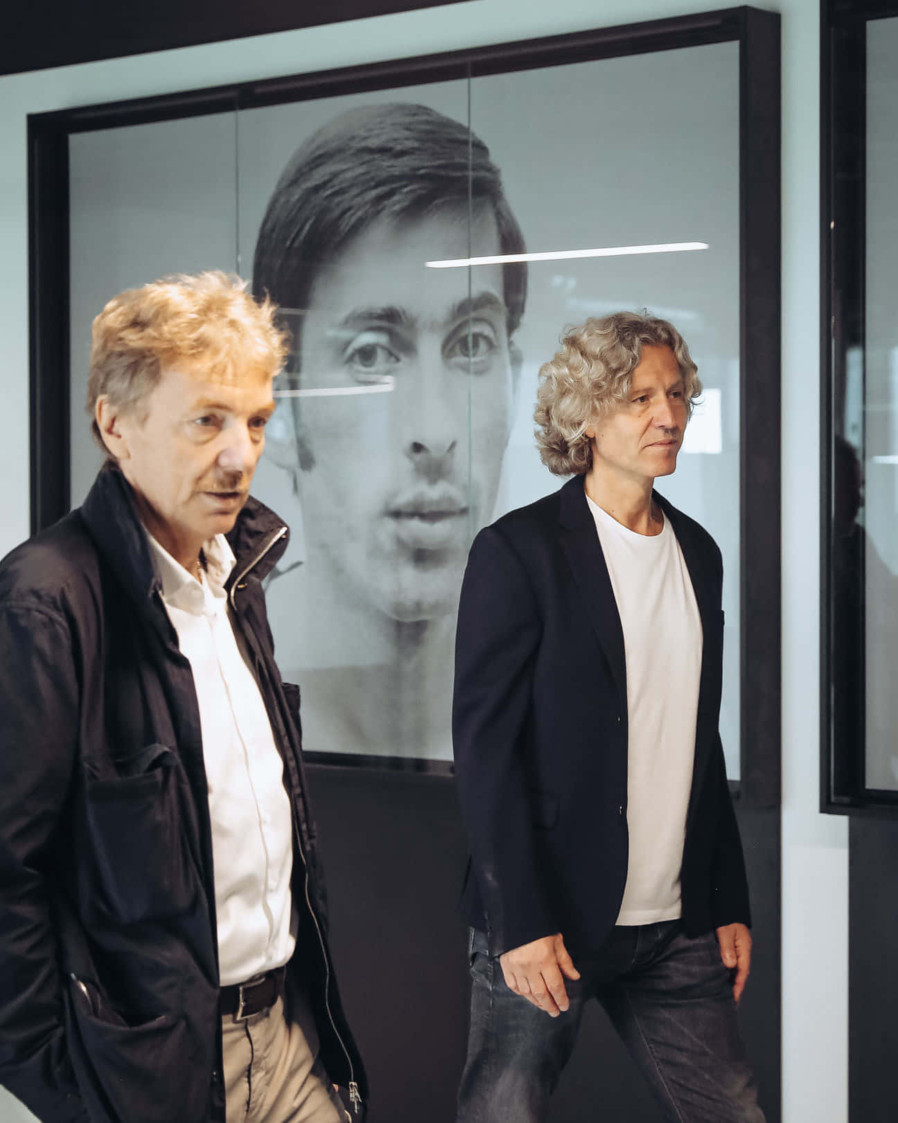 齊格尼·博尼克造訪列日訓練中心 壁紙 — 兩名身穿外套的男子站在現代畫廊空間中，一幅大型裱框黑白年輕男子肖像前。