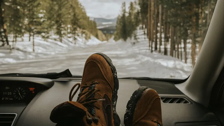 棕色登山靴擱在汽車儀表板上,面向一條積雪的森林道路。