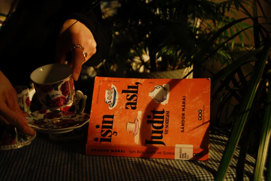 一隻手拿著一個花卉圖案的茶杯,旁邊放著一本書名為 Işın Aslı Judit 的書。