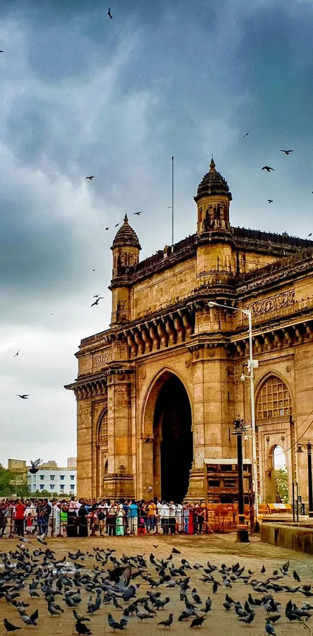 Gateway of India monument with pigeons and tourists under cloudy sky.