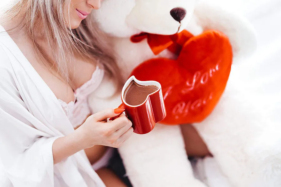 身穿白色浴袍的女性,手持紅色心形杯,旁邊有泰迪熊和「I Love You」枕頭。