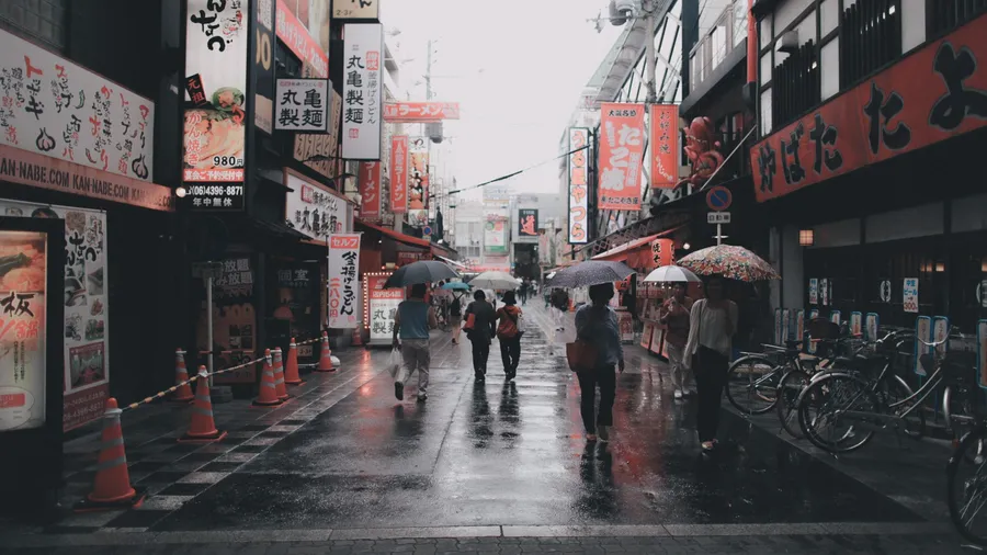 人們撐著傘走在雨中的日本街道上,兩旁有霓虹招牌和單車。