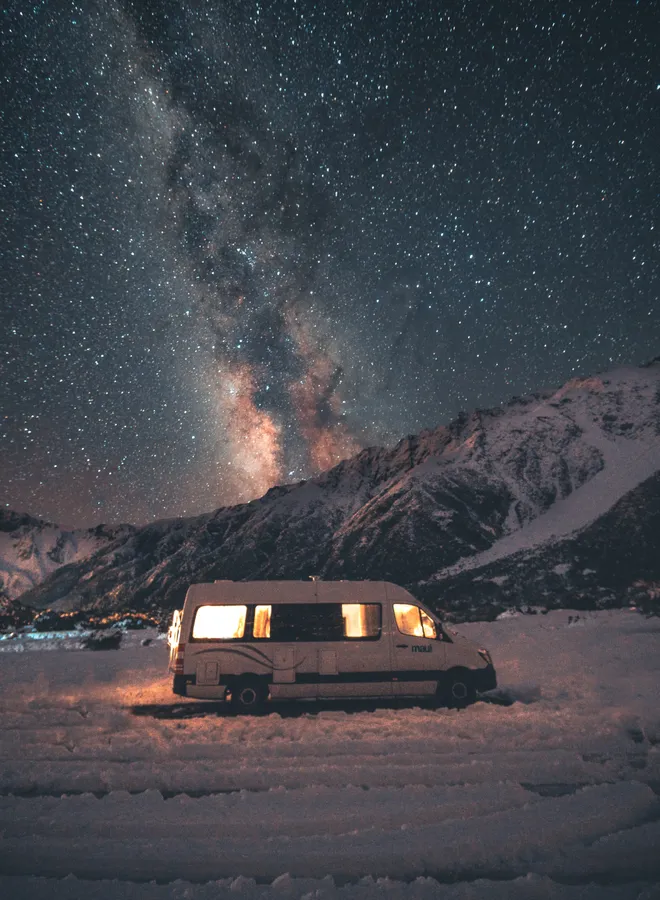 一輛露營車停在雪地中,窗戶在星空和銀河下發光。