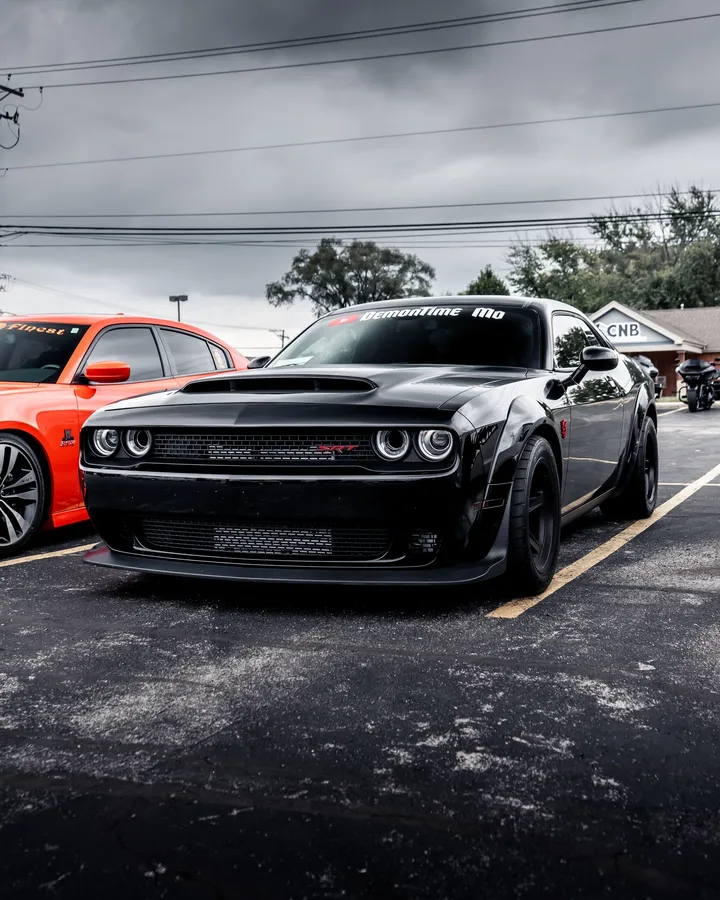 黑色Dodge Challenger Hellcat停泊在停車場中橙色汽車旁邊,天空灰暗。
