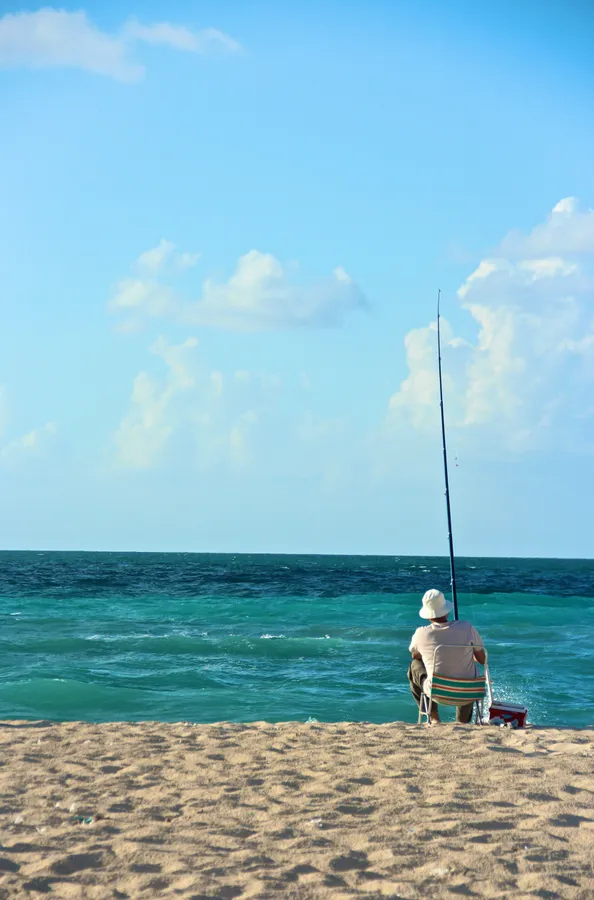 一位年長男子在陽光明媚的海灘上獨自釣魚,海水碧綠,天空湛藍。