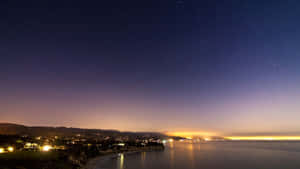 夜晚的沿海小鎮，星空璀璨，燈光倒映在水面上 — 日落天空 壁紙