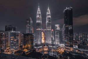 Nighttime cityscape featuring iconic twin towers and surrounding skyscrapers lit up with warm lights.
