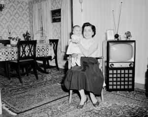 女士在1950年代客廳中抱著嬰兒，旁邊有電視和圖案牆紙。