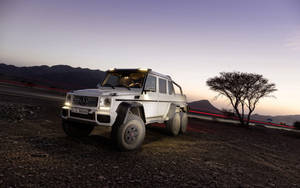 白色平治 G63 AMG 6x6 在黃昏時停在碎石上，帶有車流光跡和山巒。