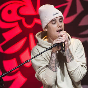 Justin Bieber 在舞台上對著麥克風唱歌，身穿米色連帽衫和白色無邊帽。