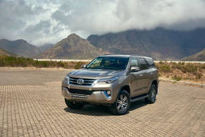 銀色豐田 Fortuner SUV 停泊在鋪砌地面上，背後有山和雲。