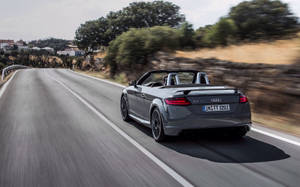 灰色Audi TT Roadster在彎曲的鄉村道路上快速行駛，背景有樹木和房屋。