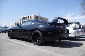 黑色豐田Supra，尾翼升起，停泊在陽光普照的城市停車場。