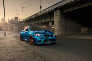 日落時分，一輛藍色BMW M2雙門跑車配備銅色輪圈，停泊在高架橋下。