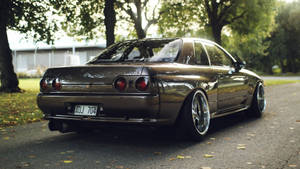 一輛青銅色日產Skyline R32配備定制輪圈，在日落時分停泊在綠樹成蔭的道路上。