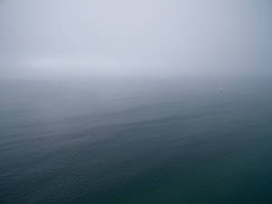 一艘小帆船在霧氣瀰漫的海洋上，帶有柔和的藍色和灰色調。 — 4K 海灘壁紙