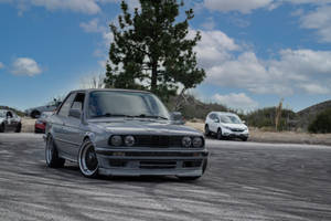 銀色BMW E30雙門轎跑車停泊在泥路上，背景有其他車輛和樹木。
