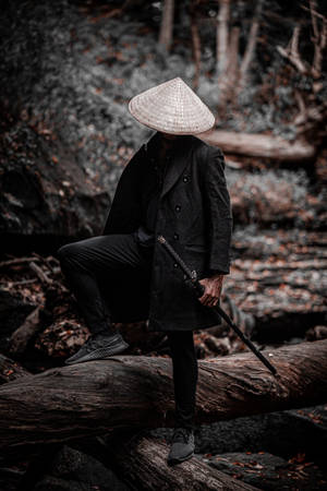 一個戴著圓錐形帽子的斗篷人物坐在森林中的一根倒下的木頭上，手持一把劍。 — 日本武士 壁紙