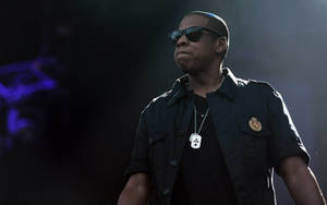 JAY Z 在舞台上表演，戴著太陽眼鏡和穿著帶有狗牌的黑色襯衫。