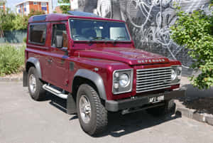 栗色Land Rover Defender停泊在陽光明媚的街道上的塗鴉牆旁。 — Surface Laptop 壁紙