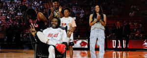 身穿白色Heat球衣的籃球運動員在球場上微笑並做出和平手勢，家人和隊友鼓掌。 — 超寬螢幕 壁紙