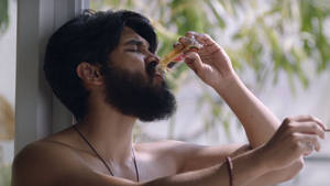 一個留鬍子的男人靠在窗邊，從玻璃瓶啜飲。