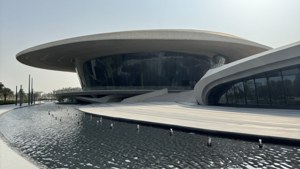 一座現代弧形建築，玻璃牆和白色屋頂，位於水景旁。