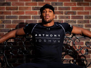 拳擊手 Anthony Joshua 身穿品牌上衣，坐在裝飾長椅上，背景是磚牆。
