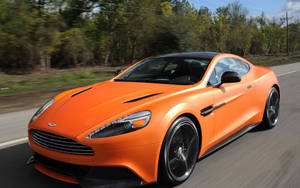 橙色Aston Martin在背景有樹木的道路上疾馳。