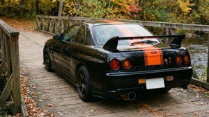 黑色日產Skyline R34帶橙色條紋停泊在秋林中的木橋上。