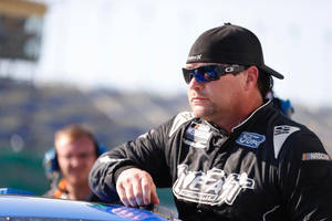 身穿黑色制服的NASCAR車手在賽道上靠在藍色賽車上。 — 黑色動態圖 壁紙