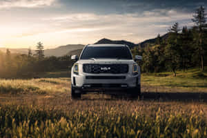 白色Kia Telluride SUV在日落時分停放在草地上，後面有山脈和樹木。 — Surface Laptop 壁紙