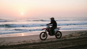 一個人騎著紅色越野電單車在日落時分沿著海灘行駛，伴隨著海浪。
