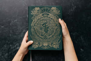 兩隻手捧著一本綠色皮革書，帶有金色龍和十字架圖案。
