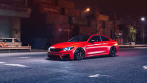 夜間濕潤的城市街道上停放的紅色BMW M4，車頭燈閃爍，後方有堆疊的貨運集裝箱。