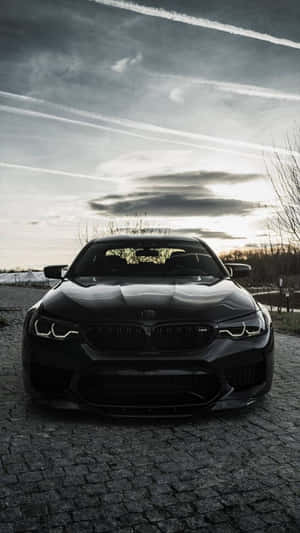 夕陽下鵝卵石路上的黑色BMW M5，天空戲劇性。 — 寶馬 iPhone 壁紙
