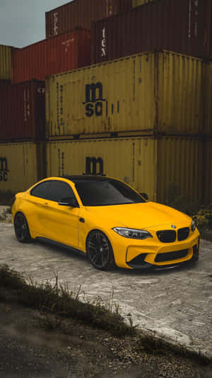 工業環境中，一輛黃色BMW M2雙門跑車停泊在堆疊的貨運集裝箱旁。