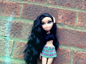 穿著格子裙的 Bratz 公仔靠在帶有綠色塗鴉的磚牆上。