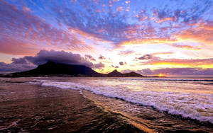 夕陽下的桌山，海浪在多彩雲層下拍打沙灘。