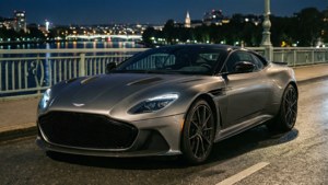 銀色Aston Martin DB11停泊在濕滑的橋上，夜晚城市燈光倒映在水面。